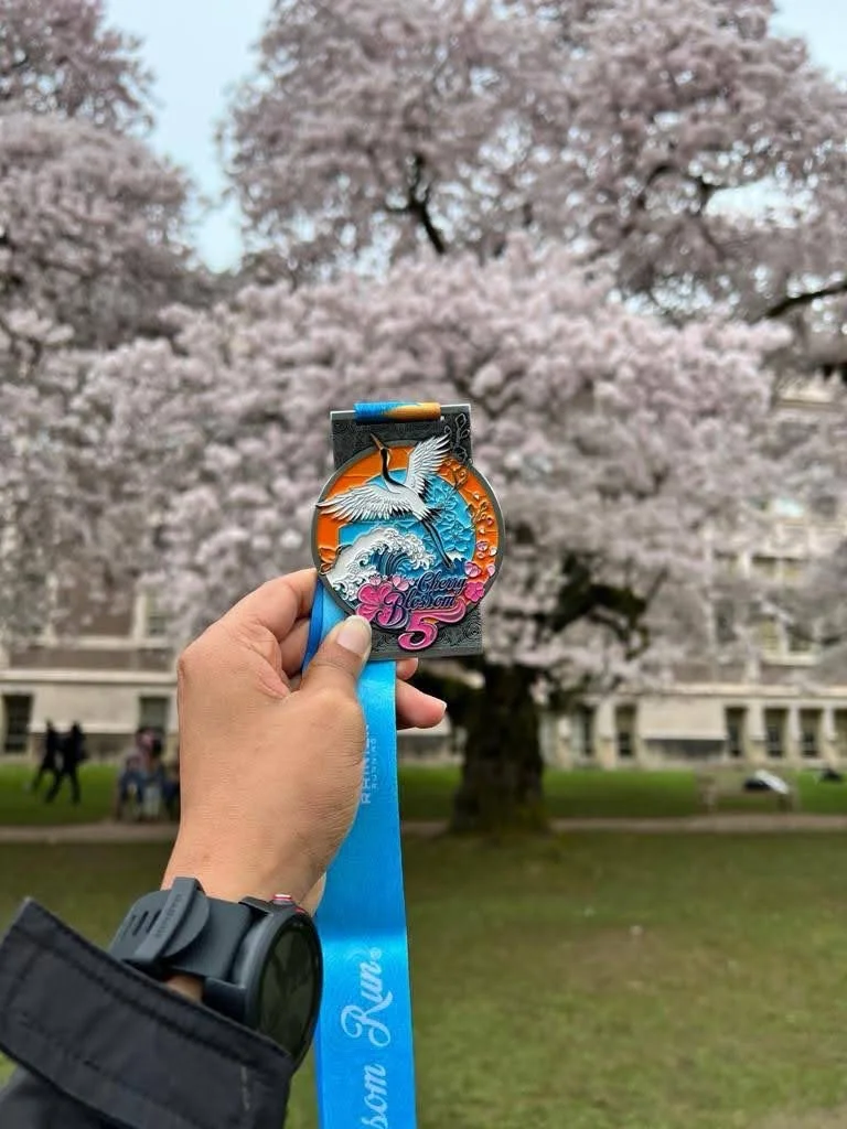 Karthika holding a Cherry Blossom 5K finisher medal under blooming cherry trees