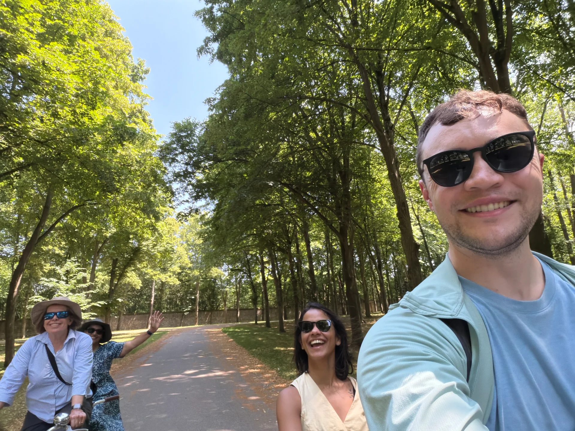 Selfie tandem biking down a tree-lined path in the Versailles gardens, the moms waving behind